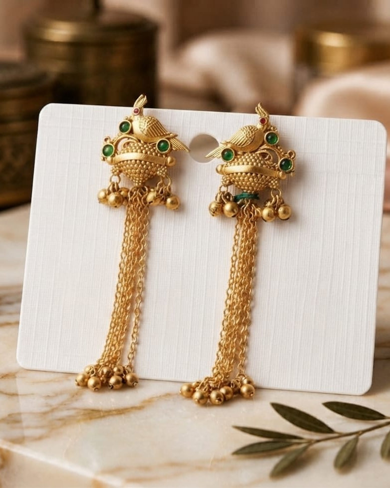 A pair of gold-toned traditional Indian Kundan earrings with green stone accents and dangling chain tassels, displayed on a white card over a marble surface with soft-focus decorative background elements.