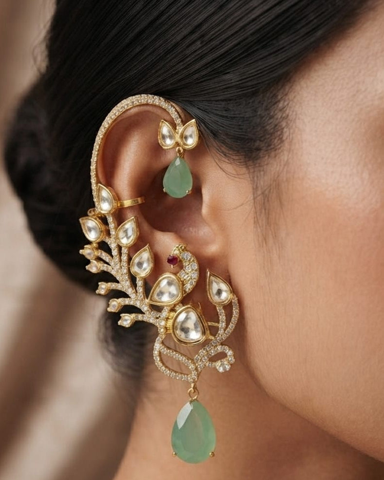 A close-up of a woman's ear wearing an ornate gold ear cuff decorated with clear stones and a green teardrop gemstone, alongside a matching small green teardrop earring.