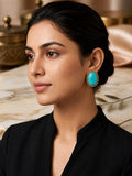 A woman with dark hair pulled back wears a large oval turquoise earring with a gold-toned textured border, paired with a black top. The background features a soft-focus warm interior with marble and gold-toned decor.