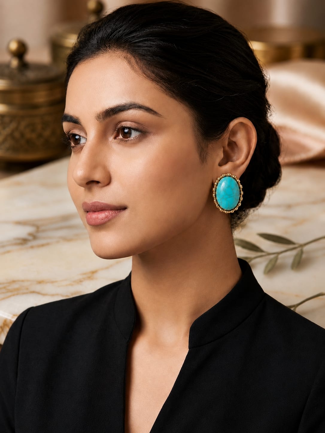 A woman with dark hair pulled back wears a large oval turquoise earring with a gold-toned textured border, paired with a black top. The background features a soft-focus warm interior with marble and gold-toned decor.
