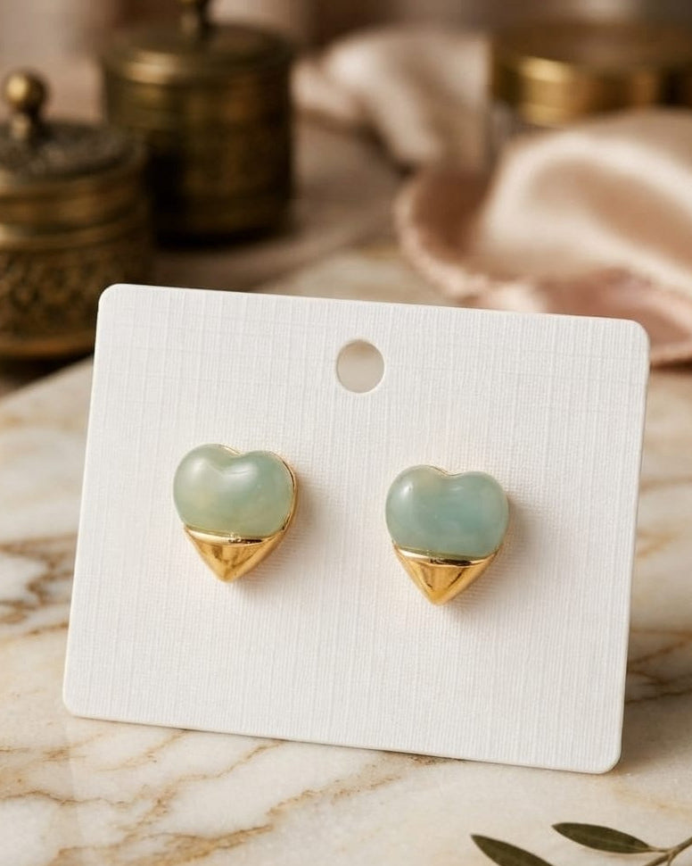 Heart-shaped stud earrings with pale green stone tops and gold triangular bases, displayed on a white card stand over a marble surface with soft decorative props in the background.