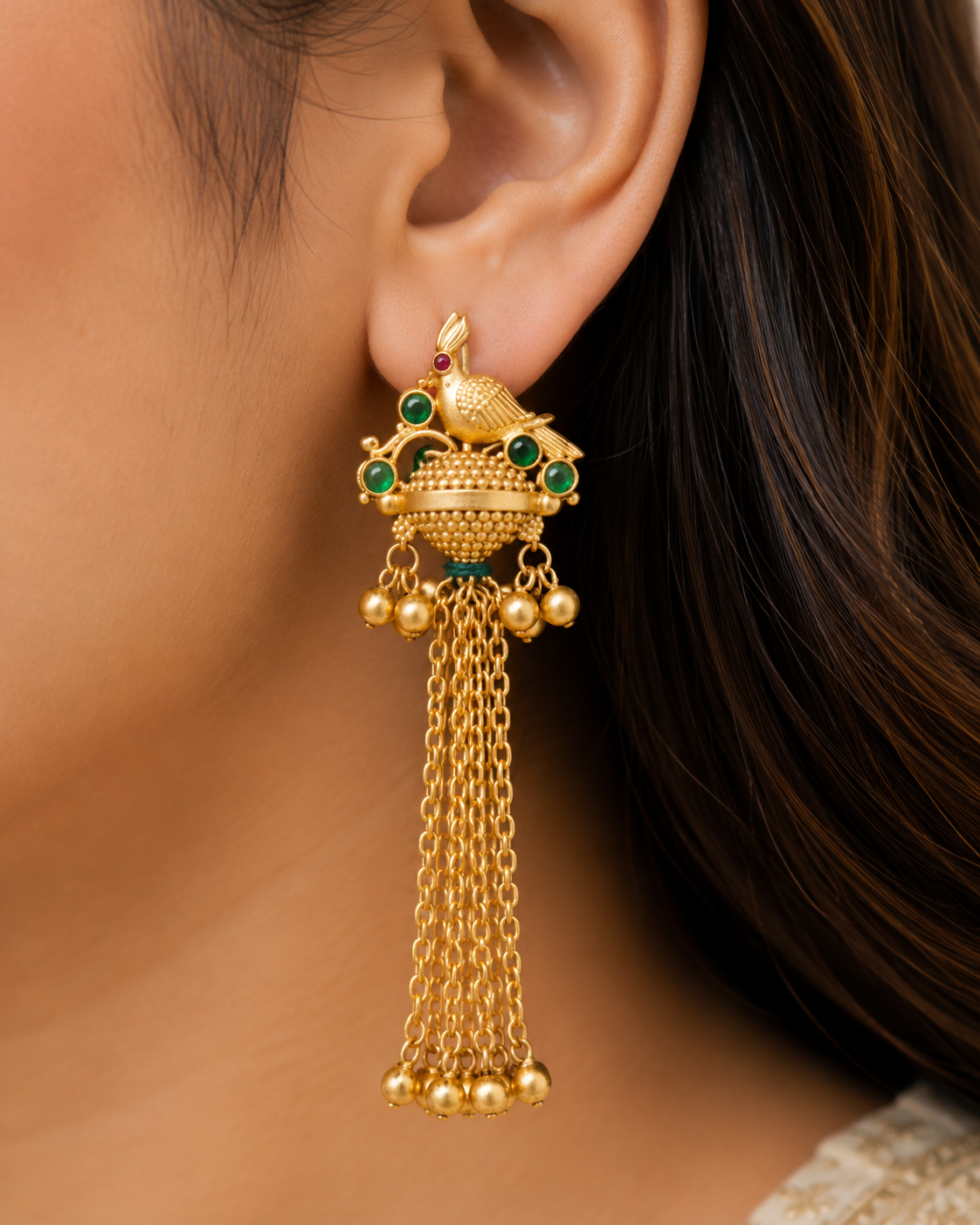 A close-up of a woman's ear wearing a traditional gold-toned Indian jhumka earring with green stone accents and dangling chain tassels.