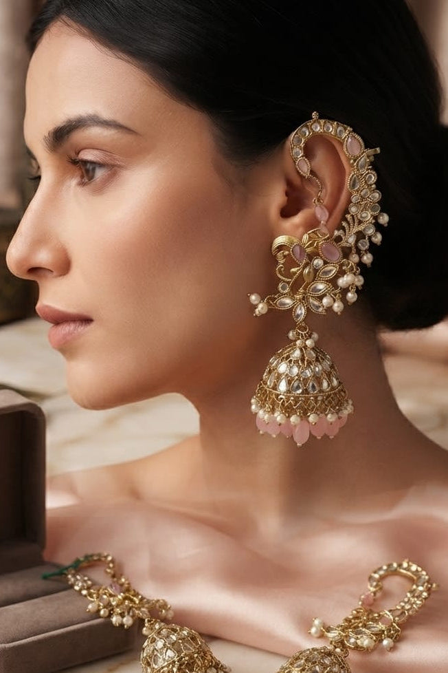 A woman in profile wearing ornate gold-toned traditional earrings with dangling bell-shaped charms and pink stone accents, with matching earrings displayed on a marble surface beside an open jewelry box.