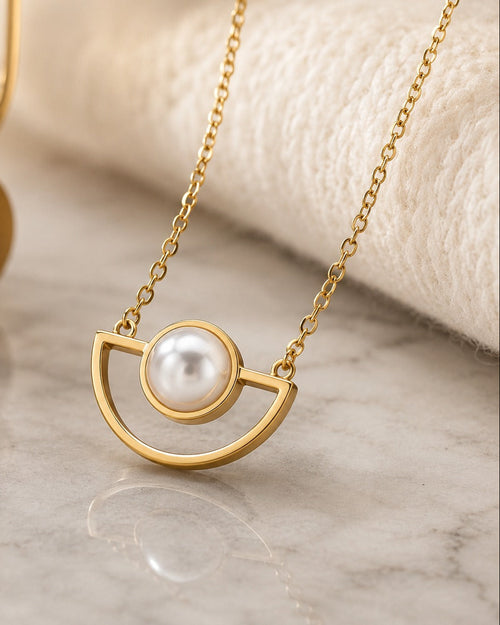 Gold necklace with a semi-circular open pendant and a central white pearl, displayed on a light marble surface with a soft cream fabric backdrop.