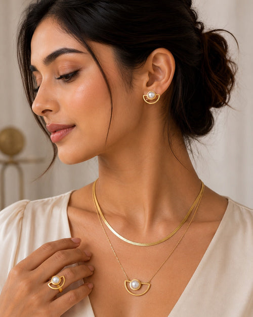 A woman wearing a gold jewelry set featuring a crescent moon pendant necklace with a white pearl, a matching thin chain necklace, and a matching crescent moon stud earring.