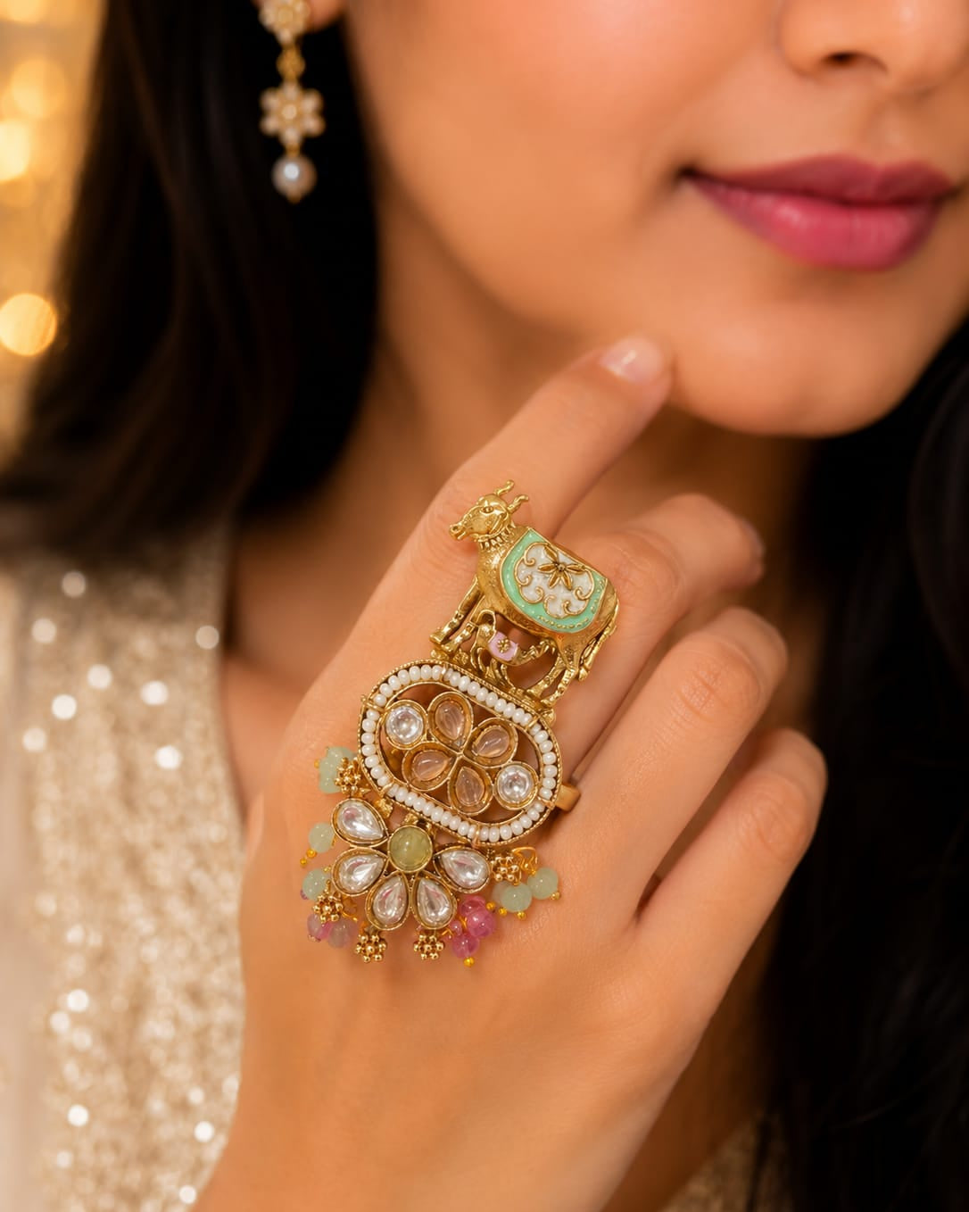 A close-up of a woman's hand wearing an ornate gold ring decorated with white pearls, green enamel, and pink stones, set against a warm, softly blurred background.