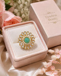 A gold ring with a round green cabochon and clear rhinestone accents, displayed in a pink velvet ring box next to its pink packaging box labeled 'Timeless Beauty'.