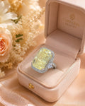 A large rectangular yellow gemstone ring with a silver halo setting, displayed inside an open beige jewelry box. The box interior is branded 'TIMELESS BEAUTY', and soft peach flowers and draped satin fabric surround the box.