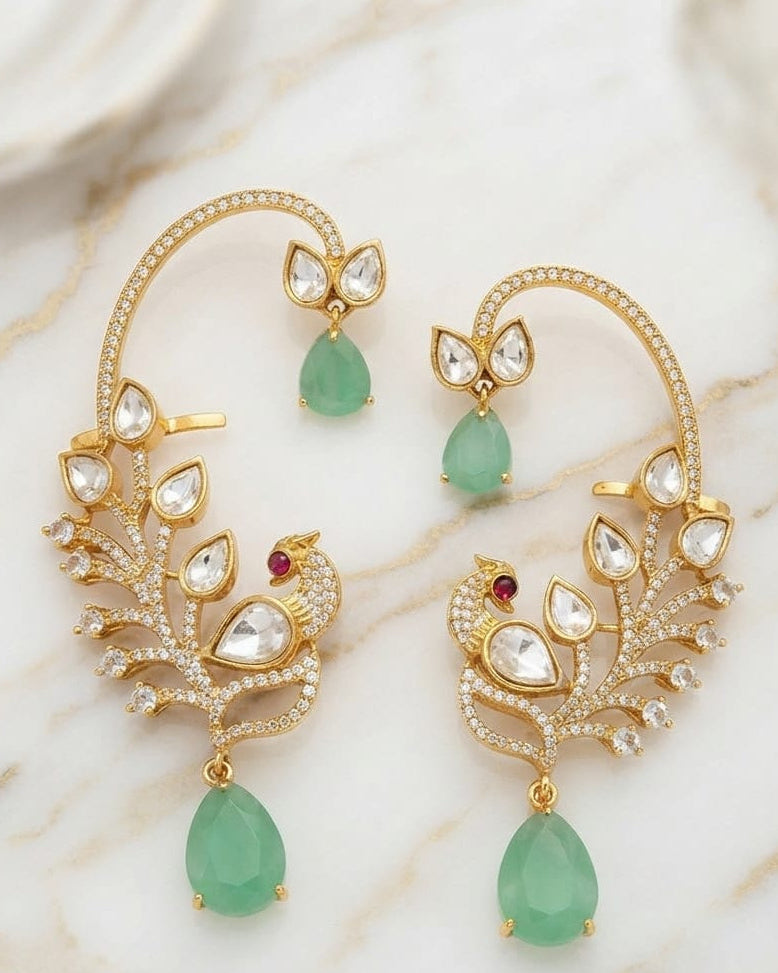 A pair of gold crescent-shaped huggie earrings decorated with clear pavé stones, small red accent stones, and green teardrop gemstones, displayed on a white marble surface.