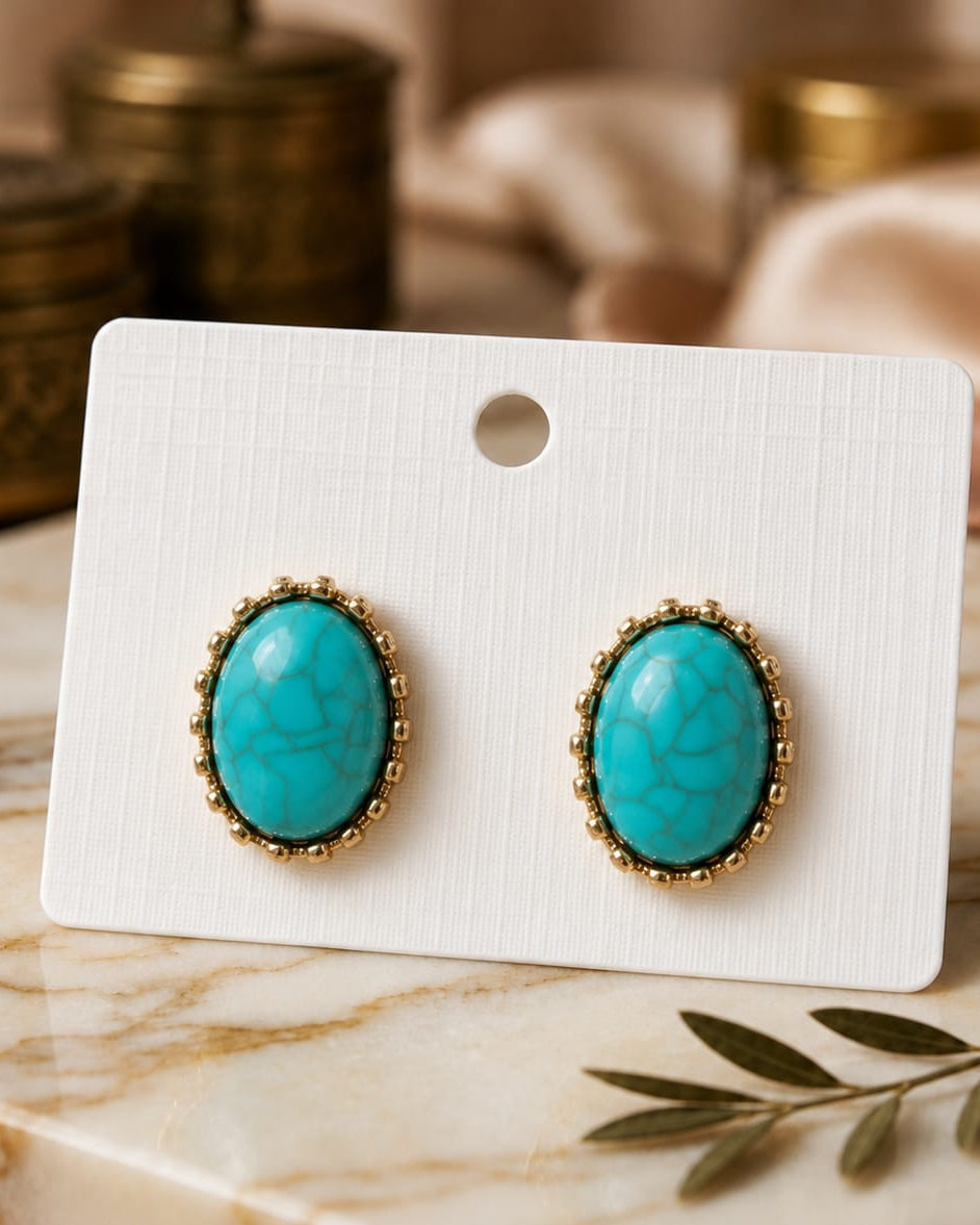 A pair of oval turquoise stud earrings with gold beaded borders, displayed on a white card stand on a marble surface with a soft-focus background.