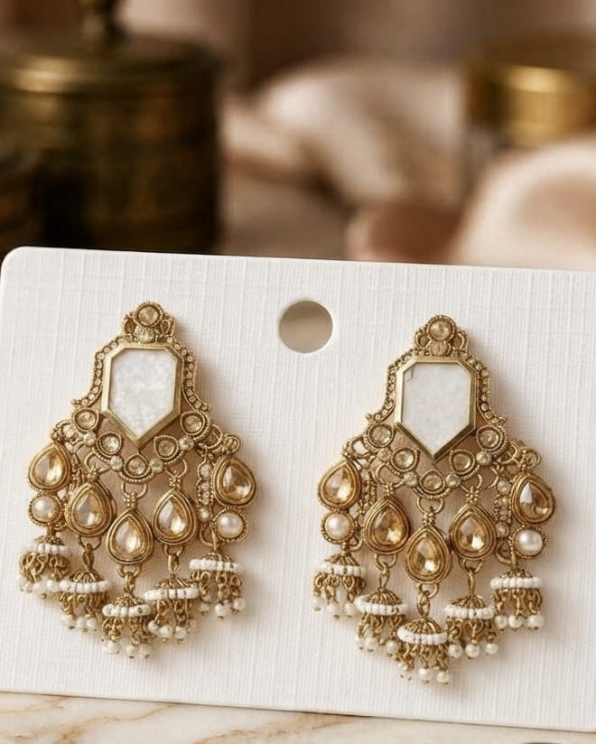 A pair of ornate gold-toned chandelier earrings with white faux pearls, displayed on a white card against a warm, softly blurred background with brass containers and a marble surface.