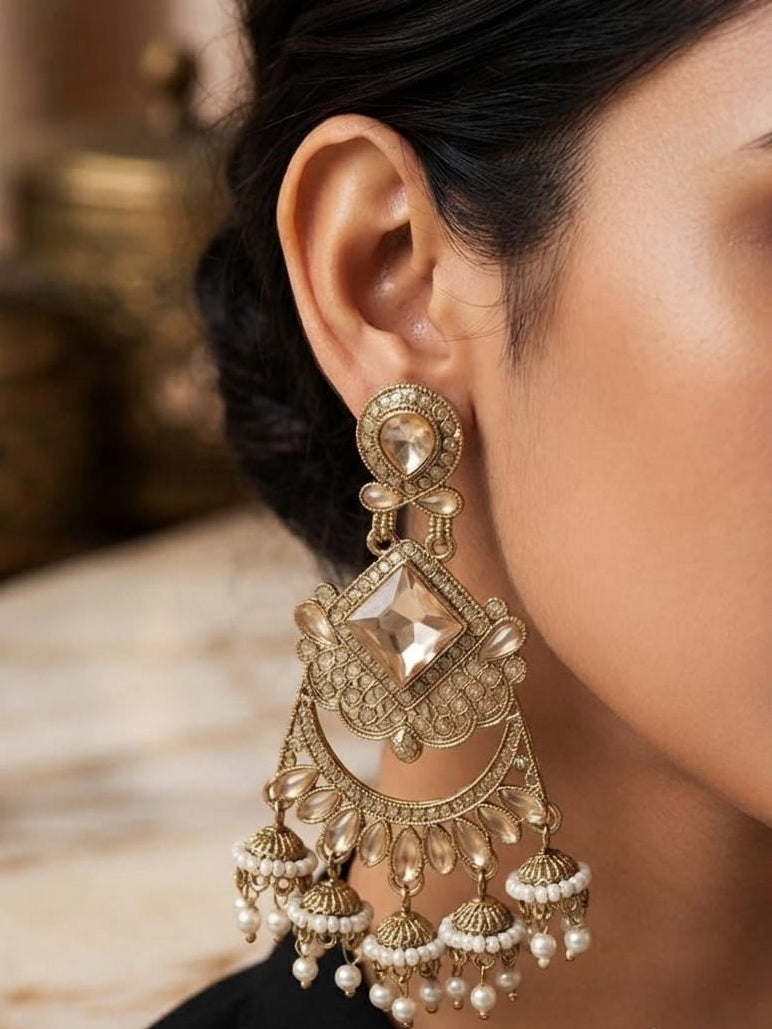 A close-up of a woman's ear wearing a large, ornate gold-toned traditional Indian chandelier earring with a central clear stone and dangling pearl-like beads.