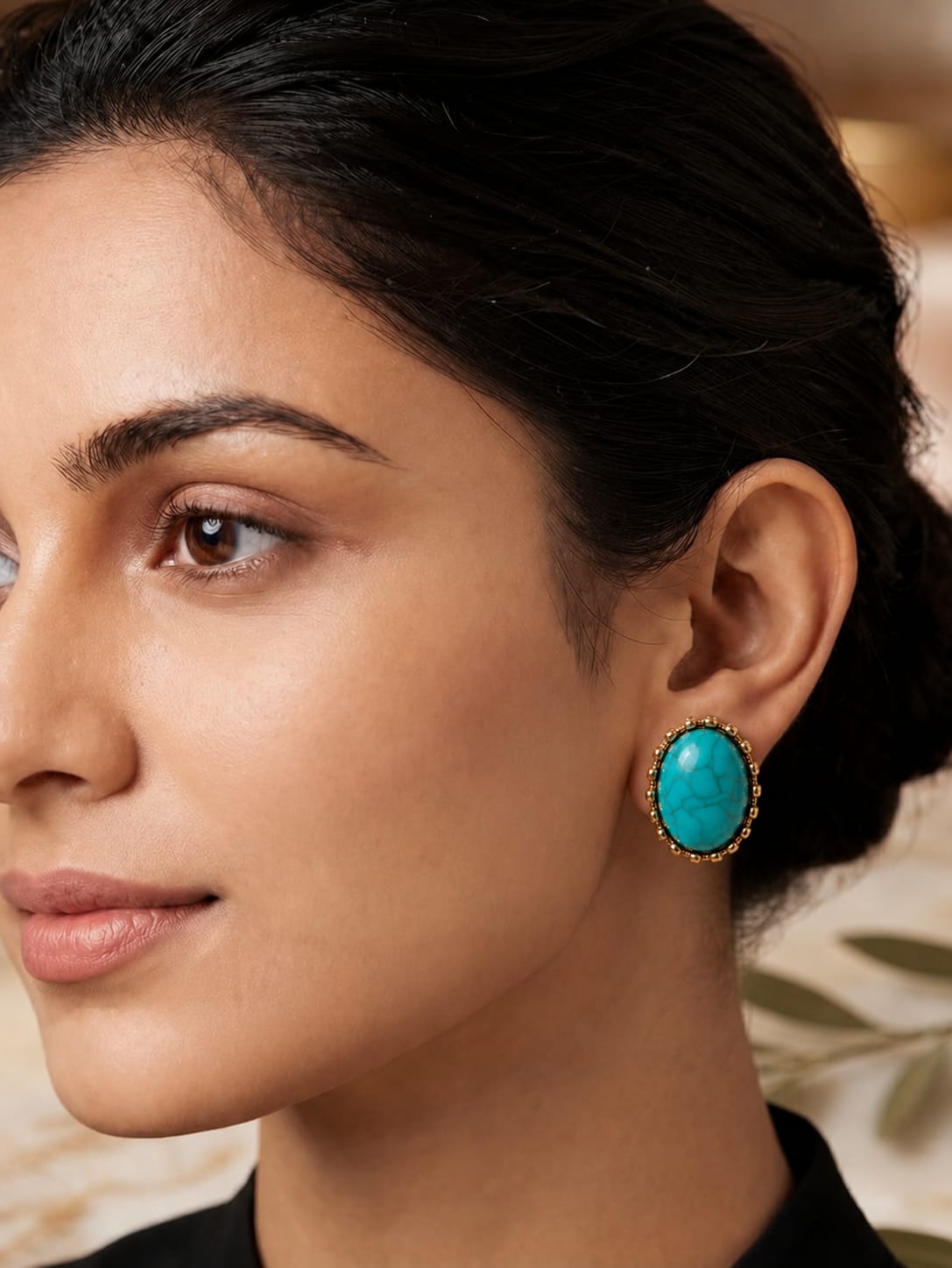 A close-up of a woman wearing a single oval turquoise earring with a gold-toned halo setting.