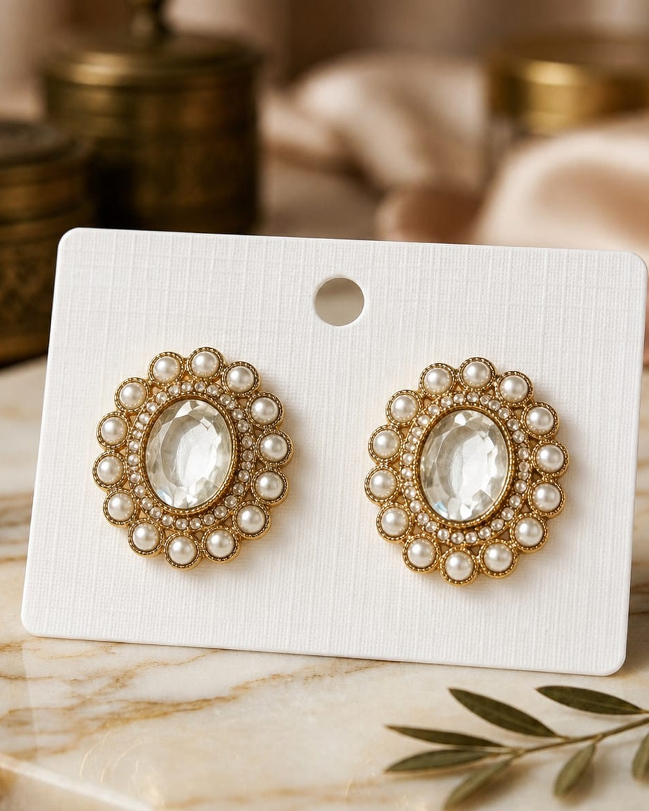 A pair of ornate oval stud earrings with clear oval center stones and rows of small white pearls around gold-tone borders, displayed on a white card against a softly blurred vintage-style tabletop background.