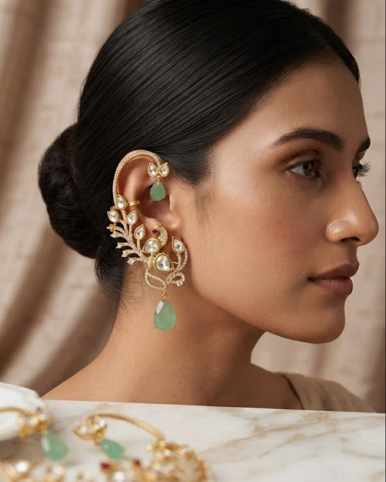 A woman in profile wearing an ornate gold-toned ear cuff with green teardrop stones and clear stones, with a matching gold-toned necklace resting on a marble surface in the foreground.