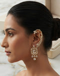 A woman in profile wearing an ornate gold-toned ear cuff and matching drop earrings with clear stones, set against a soft beige interior background.