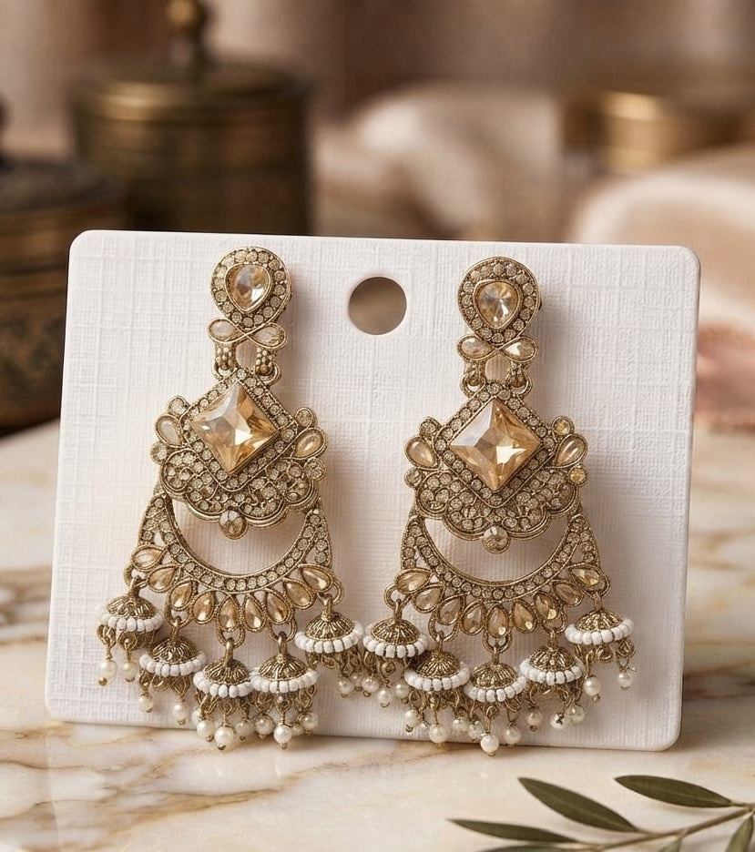 A pair of ornate gold-toned chandelier earrings with clear stones and white pearl fringe, displayed on a white card over a marble surface with a blurred decorative background.