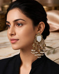 A woman with dark hair pulled back wears a prominent round gold-toned statement earring with a white pearl center and surrounding clear stones. She is wearing a black top, photographed in a warm, elegant indoor setting with marble and gold-toned decor in the background.